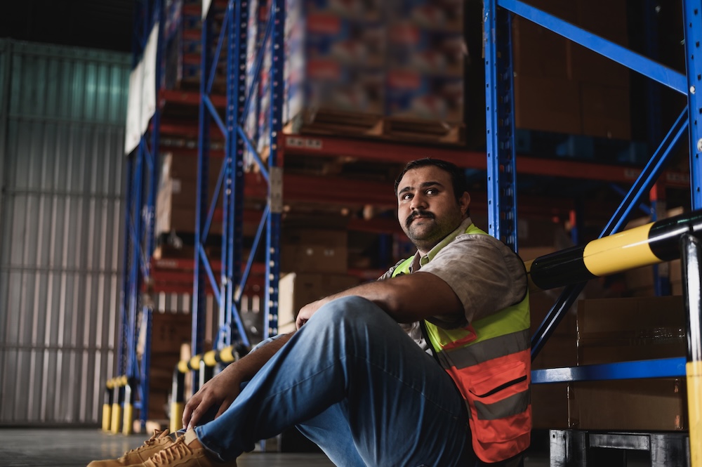 Tired warehouse worker sitting on the floor against warehouse shelves.